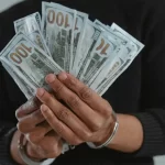 Dolandırıcılık Suçu (TCK 157) 1 Kelepçeli ellerinde dolar banknotları tutan kişi; TCK 157 dolandırıcılık suçu, hileli menfaat temini ve ceza soruşturması temasını temsil eden görsel.