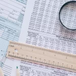 İkmalen Tarhiyat 2 İkmalen tarh işlemini anlatan görsel; vergi formları üzerinde büyüteçle inceleme ve hesaplama tablosu.