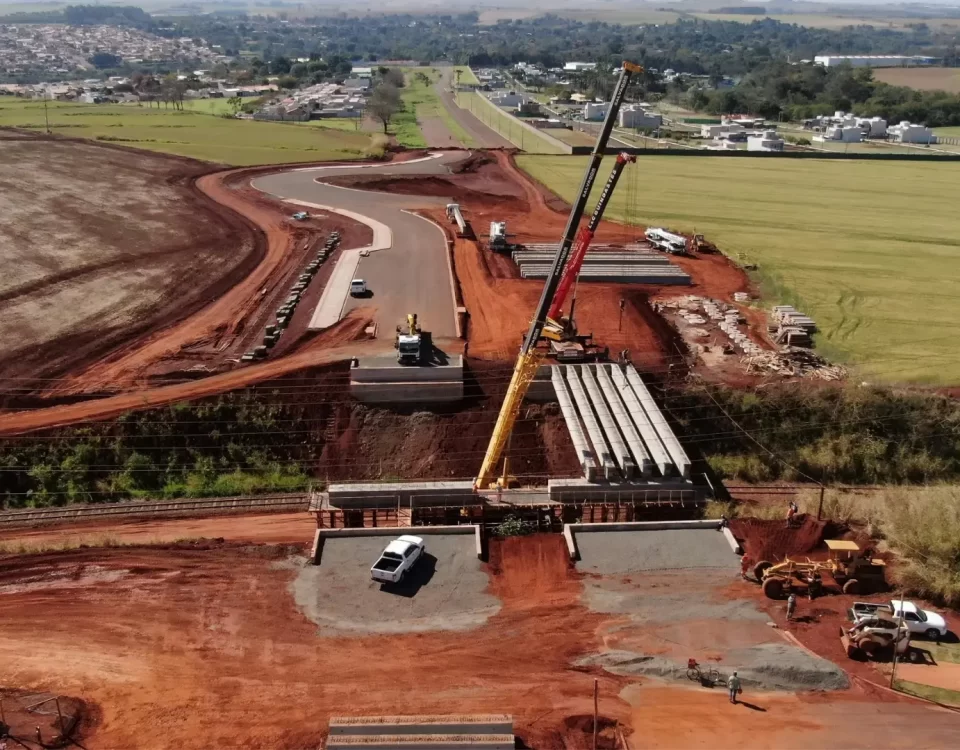 Bir yol üst geçidi inşaatının havadan görünümü. İki büyük vinç, mevcut bir tren yolunun ve tarım arazilerinin üzerinden geçen köprüye beton kirişler yerleştiriyor. Kırmızı toprak zemin ve iş makineleri görülüyor. Görsel, bir yol projesi kapsamında özel mülkiyete fiilen el atıldığı, kamulaştırmasız el atma davası senaryosunu canlandırmaktadır.