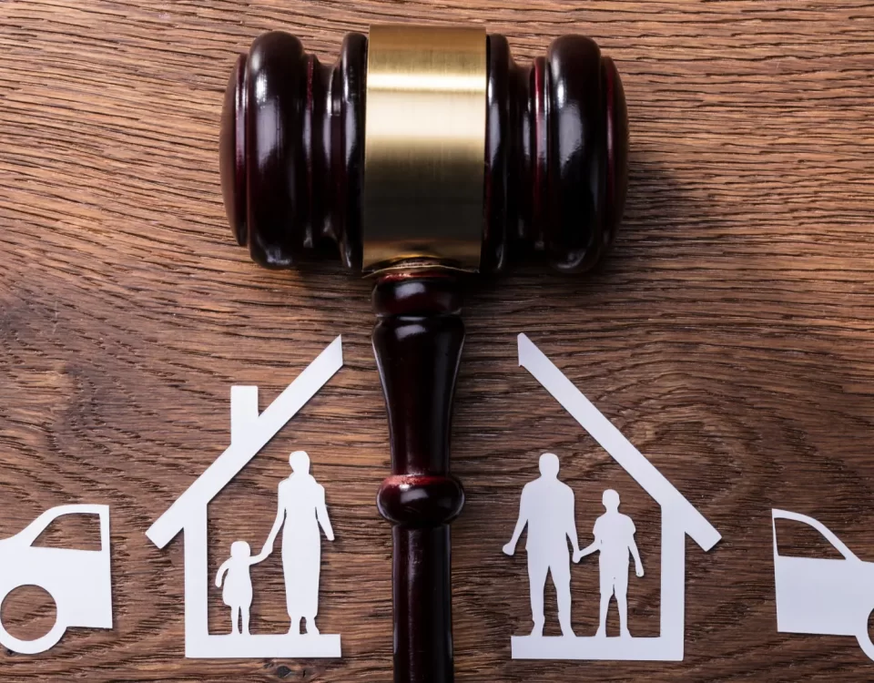 Ahşap zemin üzerinde ortadan ikiye ayrılmış kağıttan ev, araba ve aile figürleri ile adaleti simgeleyen hakim tokmağı. Görsel, boşanmada mal paylaşımı ve mal rejimi tasfiyesi davası sürecini temsil etmektedir.