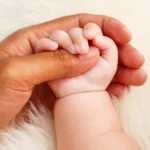 Evlat Edinme Şartları ve Süreci: Güncel Hukuki Rehber 1 Türk Medeni Kanunu uyarınca küçüklerin evlat edinilmesi sürecini, koruyucu aile ve evlat edinme şartları kapsamındaki yasal koruma bağını sembolize eden; beyaz yumuşak bir zemin üzerinde bir yetişkinin şefkatle tuttuğu bebek elini yakından gösteren kurumsal aile hukuku görseli.