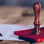 Kamu Alacaklarında İhtiyati Haciz: 6183 Sayılı Kanun Kapsamında Şartlar ve Süreç 1 6183 sayılı Amme Alacaklarının Tahsil Usulü Hakkında Kanun kapsamında vergi dairesinin uyguladığı kamu alacaklarında ihtiyati haciz kararlarını, idari merci kararlarının kesinleşmesini ve vergi mahkemelerinde açılacak iptal davalarını sembolize eden; ahşap bir masa üzerinde kırmızı renkli ıstampa pedi, dikey duran ahşap saplı kurumsal bir mühür, resmi bir hukuki evrak ve siyah renkli şık bir dolma kalemin yakın çekim görünümü.