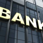 Miras Bırakanın Banka Hesaplarına Erişim: Mirasçıların Hakları ve Sınırları 2 Vefat eden murisin bankalarda bulunan mevduat, vadeli-vadesiz hesap ve kiralık kasa varlıklarının tespiti ile miras bırakanın banka hesaplarına erişim sürecini sembolize eden; modern bir finans merkezinin cam dış cephesinde yer alan büyük altın sarısı renkli "BANK" tabelasını ve gökyüzüne doğru uzanan kurumsal banka binasını gösteren mimari hukuk görseli.