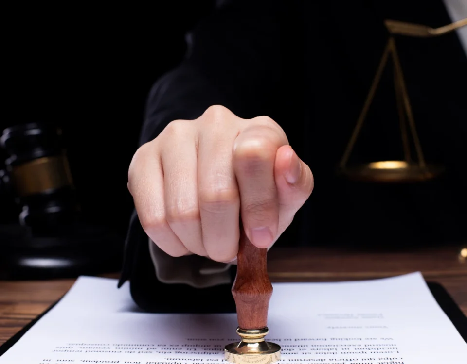 Saklı Pay Oranları ve Korunması: Tenkis Davası Rehberi 6 Türk Medeni Kanunu uyarınca mirasbırakanın tasarruf özgürlüğünü kısıtlayan ve koruma altına alınan saklı pay oranlarının ihlali durumunda açılan tenkis davası mahkeme karar sürecini sembolize eden; ahşap masa üzerindeki resmi yasal belgeye mühür/kaşe basan cübbeli bir hukukçunun elini, arka planda ise adalet terazisini ve hakim tokmağını gösteren kurumsal miras hukuku görseli.