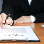 Şirkete Kayyum Atanması: Vergi Suçlarında Şartlar, Sorumluluk ve Güncel Yargı Uygulaması 2 Türk Ticaret Kanunu uyarınca ortaklar arasındaki uyuşmazlıklar veya yönetim organının işlevsiz kalması nedeniyle mahkemece verilen şirkete kayyum atanması kararını ve denetim sürecini sembolize eden; ahşap masa üzerindeki resmi şirket yönetim kurulu evraklarını ve karar metnini dolma kalemle inceleyip onaylayan, siyah takım elbiseli kurumsal bir şirketler hukuku avukatı ya da mahkemece atanan resmi kayyum figürü.