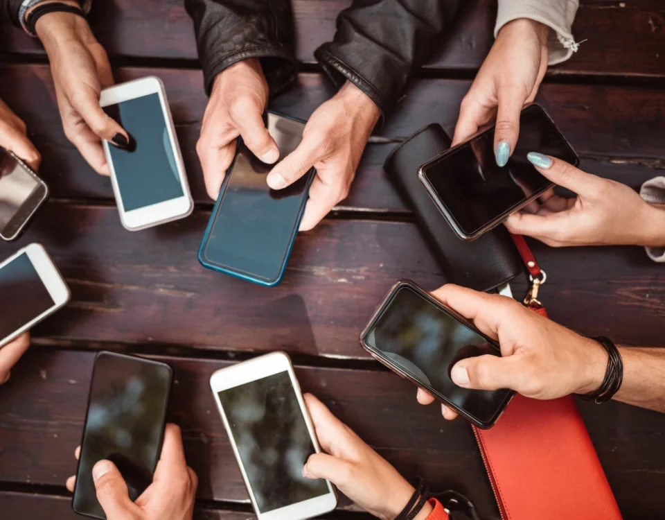 Sosyal medyada hakaret suçu teşkil eden dijital paylaşımları, yorumları, DM mesajlarını ve siber zorbalık eylemlerini akıllı telefonları üzerinden gerçekleştiren veya bu içeriklere maruz kalan kişileri, internet ortamındaki kolektif dijital etkileşimi sembolize eden, bir masa etrafında ellerinde cep telefonları olan insanların üstten görünüm kurumsal ceza hukuku görseli.