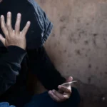 Uyuşturucu Kullanım ve Ticaret Ayrımı: TCK 188 İçin 5 Kritik Ölçüt 2 Türk Ceza Kanunu kapsamında uyuşturucu kullanım ve ticaret ayrımı hususunda Yargıtay tarafından belirlenen kriterleri ve cezai sorumluluğu sembolize eden; siyah kapüşonlu hırka giymiş, yüzünü eliyle gizleyen ve diğer elinde şeffaf küçük kilitli poşet içinde madde bulunduran, duvara yaslanmış bir şüpheliyi gösteren kurumsal ceza hukuku görseli.
