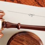 Vasiyetnamenin Tenfizi ve Açılması: TMK m. 596-602 Kapsamlı Rehber 2 Türk Medeni Kanunu uyarınca mirasbırakanın vefatı sonrası açılan ve okunan resmi vasiyetnamenin yerine getirilmesini sağlayan vasiyetnamenin tenfizi davası mahkeme aşamasını sembolize eden; fildişi rengi resmi yasal evrakların üstünde duran ahşap hakim tokmağını ve altlığının üstten görünümünü, arka planda ise kahverengi harita desenini gösteren kurumsal miras hukuku görseli.