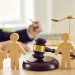 Velayetin Değiştirilmesi Davası: 2026 Kapsamlı Rehber 2 Bir hukuk ofisinde veya mahkeme salonunda, arka planda adalet terazisi ve evrak imzalayan hukukçu silüetinin önünde yer alan, ebeveyn ile çocuk ilişkilerini ve yasal ayrılık süreçlerini simgeleyen ahşap figürler ile ortadaki hâkim tokmağını gösteren, velayetin değiştirilmesi davası ve velayet hakkının kötüye kullanılması konularıyla ilgili kurumsal aile hukuku görseli.