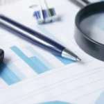 Vergi İncelemesinde Mükellef Hakları 1 Vergi Usul Kanunu kapsamında yürütülen defter ve belge denetimlerinde yasal savunma mekanizmalarını ve vergi incelemesinde mükellef hakları güvencelerini sembolize eden; üzerinde mavi renkli finansal grafik tabloları, tükenmez kalem, hesap makinesi köşesi ve denetimi temsil eden siyah çerçeveli büyüteç bulunan kurumsal mali hukuk çalışma masasının üstten yakın çekim görünümü.