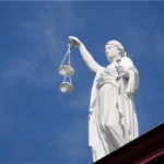 Mavi gökyüzü önünde terazi ve kılıç tutan beyaz adalet heykeli; İstanbul'da iş kazası geçiren işçilerin adliye koridorlarındaki hak arayışını ve iş kazası tazminatı davalarındaki hukuki süreci temsil eder.